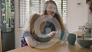 Beautiful young mother and her cute blond daughter are laughing happily at kitchen table. Slow mo