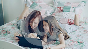 Beautiful young mother and her cute daughter in pajamas laughing and looking in digital tablet while lying on bed at