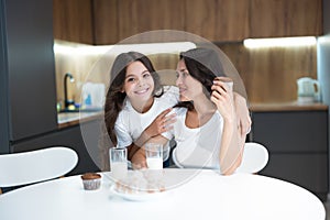 Beautiful young mother with her cute teen daughter eating cupcakes and croissants and drinking milk for breakfast in the