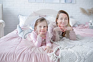 Beautiful young mother and her little daughter are lying together on the bed in the bedroom, playing, hugging and having fun.