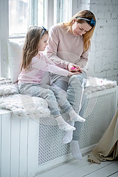 Beautiful young mother and her little daughter are sitting by the window together and painting their nails. Maternal care and love