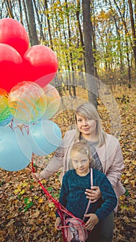 A beautiful young mother hugs her a little adorable daughter and holds the colorful balloons on a wall walk in autumn