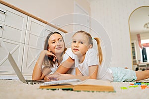 Beautiful young mother and little girl using laptop computer together lying on floor in children room with modern light