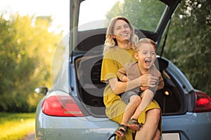 Beautiful young mother and little son travelers sit in trunk of car on road and laugh. Photo about family travel