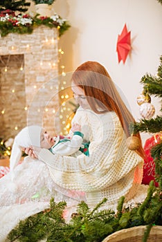 Beautiful young mother with red hair in a white knitted warm sweater holds a baby in her arms at home in a room decorated