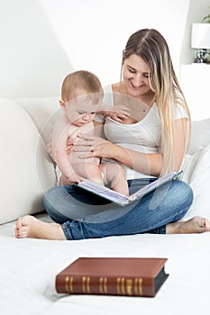 Beautiful young mother sitting on bed with her baby and reading big old book