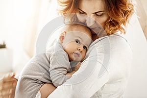 Beautiful young mother tenderly hugs her little cute child. Mom looking at her kid and smiling while baby looks aside