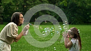 Beautiful young mother together with her daughter in nature making soap bubbles and laughing.