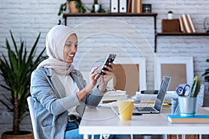 Beautiful young muslim business woman wearing hijab sending messages with mobile phone while sitting in the office