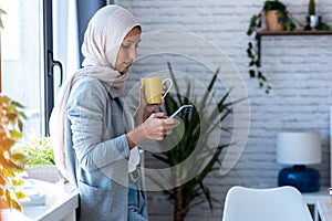 Beautiful young muslim business woman wearing hijab sending messages with mobile phone while standing near the window in the