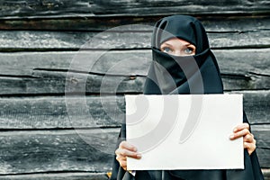 A beautiful, young, Muslim girl in a black veil with a closed face against a gray tree. In the hands of a sheet of paper. Copy