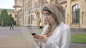 Beautiful young muslim woman in hijab typing on phone and standing on street near building, wearing white jacket