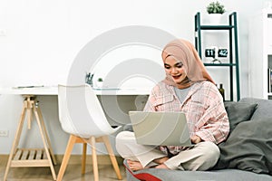 A beautiful young Muslim woman wearing casual clothing is using a laptop and smiling while working at home, sitting on a