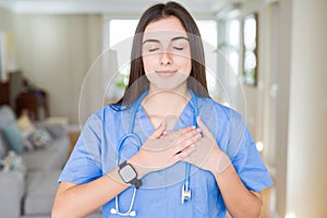 Beautiful young nurse woman wearing uniform and stethoscope at the clinic smiling with hands on chest with closed eyes and