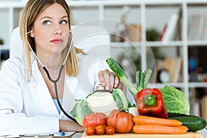 Beautiful young nutritionist listening with stethoscope on vegetables in the consultation.