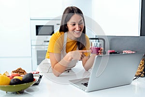 Beautiful young nutritionist woman having an online video call via laptop computer with a friend in the kitchen at home