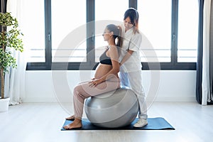 Beautiful young physiotherapist helping to beautiful pregnant woman for doing pilates exercises with ball preparing for childbirth