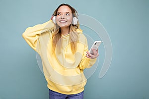 Beautiful young positive blonde woman wearing yellow stylish hoodie isolated over blue background wearing white