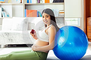 Beautiful young pregnant asian woman holding apple and lean against exercise ball in living room at house with smiley face and