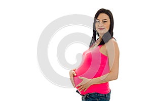 Beautiful young pregnant brunette woman in pink shirt touching her belly and looking at the camera isolated on white