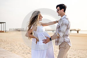 Beautiful young pregnant couple embracing and looking to each other on the beach at sunset.