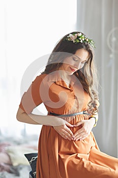 A beautiful young pregnant girl with a wreath of flowers on her head in a brown dress with lace is sitting near the window and