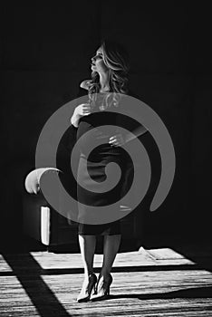 Beautiful young pregnant woman with curls and make-up in a black tight-fitting dress. Black and white photo. Fine art.