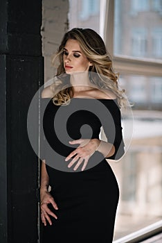 Beautiful young pregnant woman with curls and make-up in a black tight-fitting dress. Expectation of a child of 9 months.