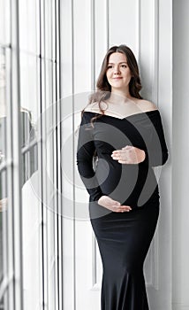 beautiful young pregnant woman in dark black dress stands near window