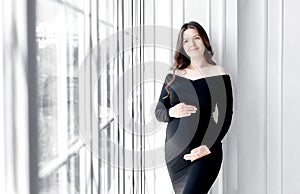 beautiful young pregnant woman in dark black dress stands near window