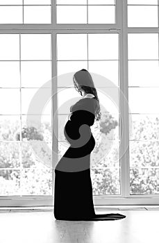 beautiful young pregnant woman in dark black dress stands near the window black and white photo