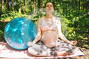 Beautiful young pregnant woman doing exercising with fitness pilates blue ball in park outdoor. Sitting and relaxing on pink yoga