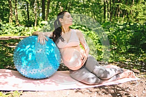 Beautiful young pregnant woman doing exercising with fitness pilates blue ball in park outdoor. Sitting and relaxing on pink yoga