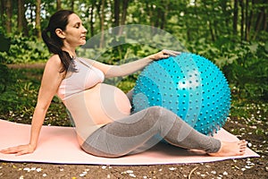 Beautiful young pregnant woman doing exercising with fitness pilates blue ball in park outdoor. Sitting and relaxing on pink yoga