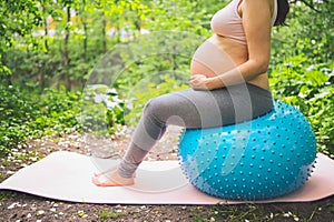 Beautiful young pregnant woman doing exercising with fitness pilates blue ball in park outdoor. Sitting and relaxing on pink yoga