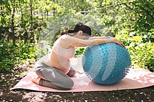 Beautiful young pregnant woman doing exercising with fitness pilates blue ball in park outdoor. Sitting and relaxing on pink yoga