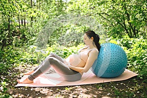 Beautiful young pregnant woman doing exercising with fitness pilates blue ball in park outdoor. Sitting and relaxing on pink yoga