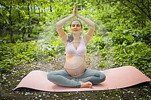 Beautiful young pregnant woman doing yoga exercising in park outdoor. Sitting and relaxing on pink yoga mat. Active future mother