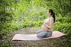 Beautiful young pregnant woman doing yoga exercising in park outdoor. Sitting and relaxing on pink yoga mat. Active future mother