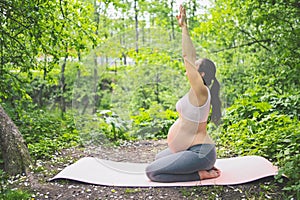 Beautiful young pregnant woman doing yoga exercising in park outdoor. Sitting and relaxing on pink yoga mat. Active future mother