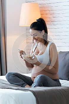 Beautiful young pregnant woman eating cereals flakes while sitting on the bed at home