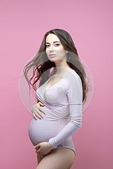 Beautiful young pregnant woman with flowing long wavy hair stands on an isolated pink background and hugs her tummy