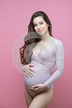 Beautiful young pregnant woman with flowing long wavy hair stands on an isolated pink background and hugs her tummy