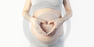 Beautiful young pregnant woman holding hands symbol heart on her belly on white background. The concept of expectation of the