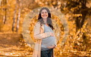 Beautiful young pregnant woman hugging her belly on walk in autumn park