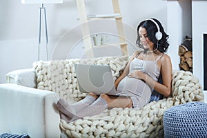 Beautiful young pregnant woman with laptop sitting on sofa at home