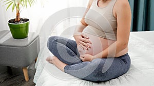 Beautiful young pregnant woman in leggings sitting on bed side and stroking her big belly against bright morning sun