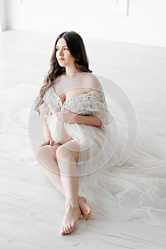 beautiful young pregnant woman in long white dress and sitting on floor