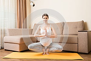 Beautiful young pregnant woman sitting on mat in yoga pose at the home. Pregnancy Yoga and Fitness concept at coronavirus time