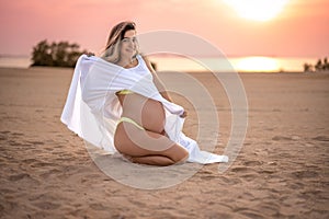 Beautiful young pregnant woman sitting on sand, holding her white dress and enjoying pregnancy on the beach at sunset.